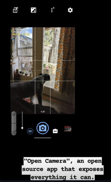 Black background.
Screenshot towards the top left corner of using a dark themed camera app (black background). Four top bar icons, mostly in white: Open padlock with small square photo contrast symbol to the top right. Square photo contrast symbol, which is split diagonally with the top left side filled in with white except a black - and the bottom right filled in with black except a white plus, as well as outlined in white. Three vertical more menu dots with a flash symbols slashed out to the top right. Settings cog.
Camera view is of a dog looking out the door window, which has a curtain covered with sunflowers pushed to the side. By the door is a window and 5 plants on the window sill. The view outside is of various plants in planters against a wooden fence and trees. There is an overlaid 3x3 grid with the dividing lines closer together to the center. In the top left: white text: 1:10:28 p.m.
29.0 GB
Yellow text: ISO 44 1/534s
Small white text: DRD.
Bottom icons: A zoom bar with a - magnifying lens on the bottom and a + magnifying lens on the top. The slider bar next to this label is has the slider dot all the way at the bottom. Blue video recording icon surrounded by circle. Big blue camera icon surrounded by circle, indicating that the user is taking a photo. Small white camera icon labelled with a black double ended horizontal arrow. Thumbnail of a dog resting with their belly on the ground.
Text annotation towards bottom right corner: "Open Camera", an open source app that exposes everything it can.
