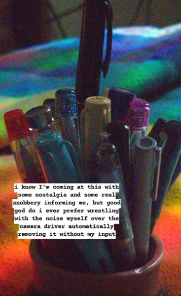 Grainy picture of full pen holder on a rainbow blanket. One black capped pen is sticking out from the bunch.
Text annotation towards bottom left: i know I'm coming at this with some nostalgia and some real snobbery informing me, but good god do i ever prefer wrestling with the noise myself over the camera driver automatically removing it without my input.